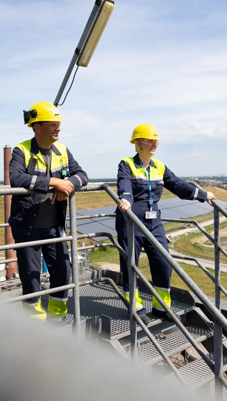 Two Oleon employees in navy overalls, high-visibility jackets and yellow hard hats stand at the railing of an elevated walkway, looking out over the solar array and fields beyond the plant.