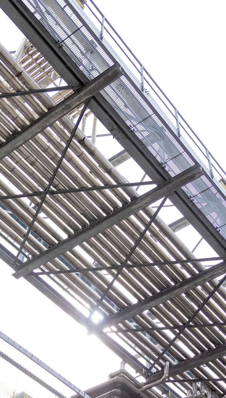 An overhead pipe bridge seen from directly beneath, its parallel runs and grated walkway cutting a diagonal across a bright white sky.